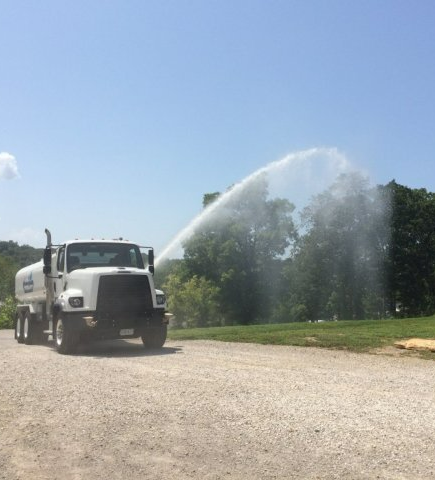 White water truck spraying a high arc of water on a gravel road beside green trees under a blue sky