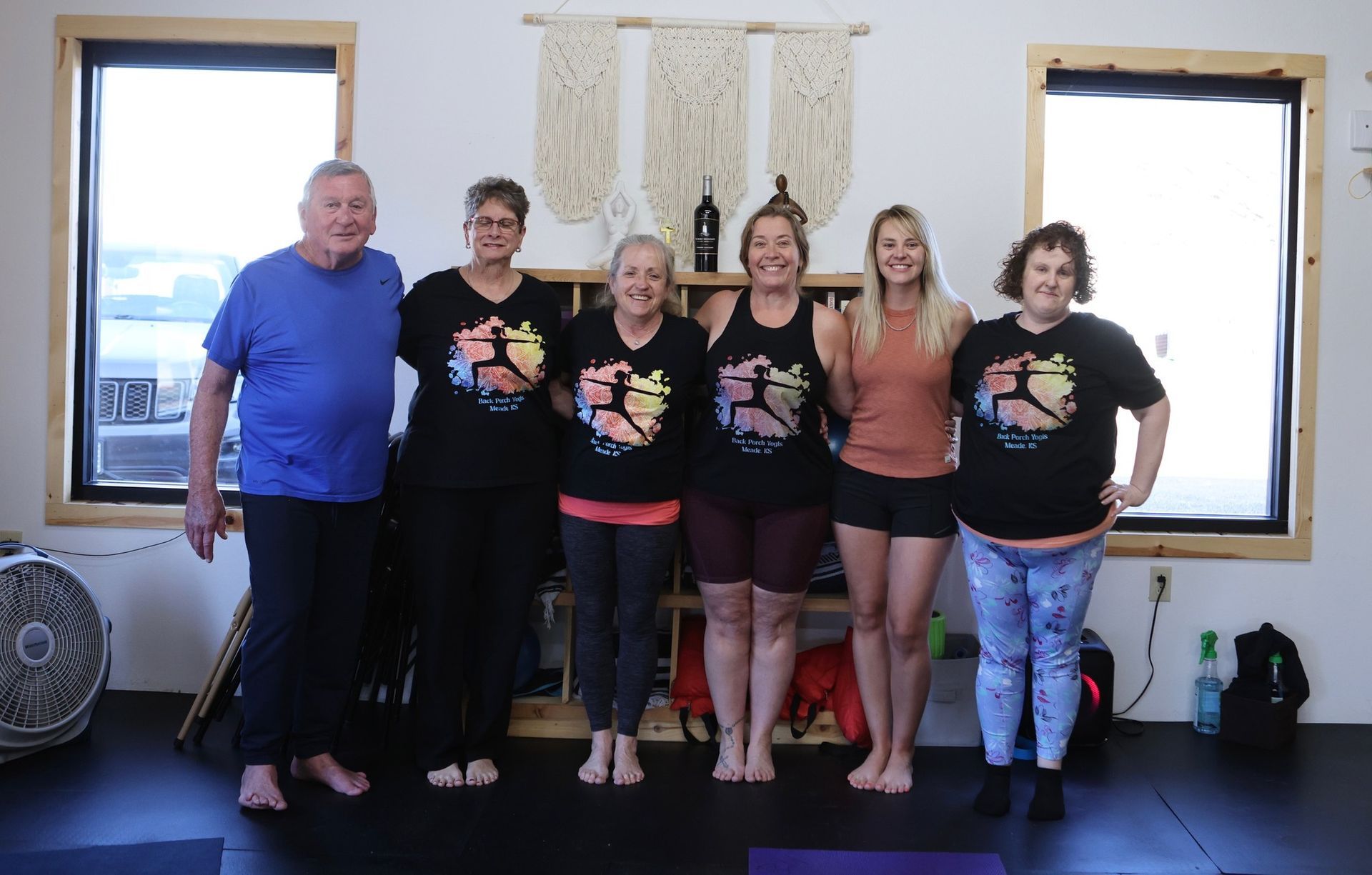 Six people posing indoors between two windows, smiling and standing arm in arm.