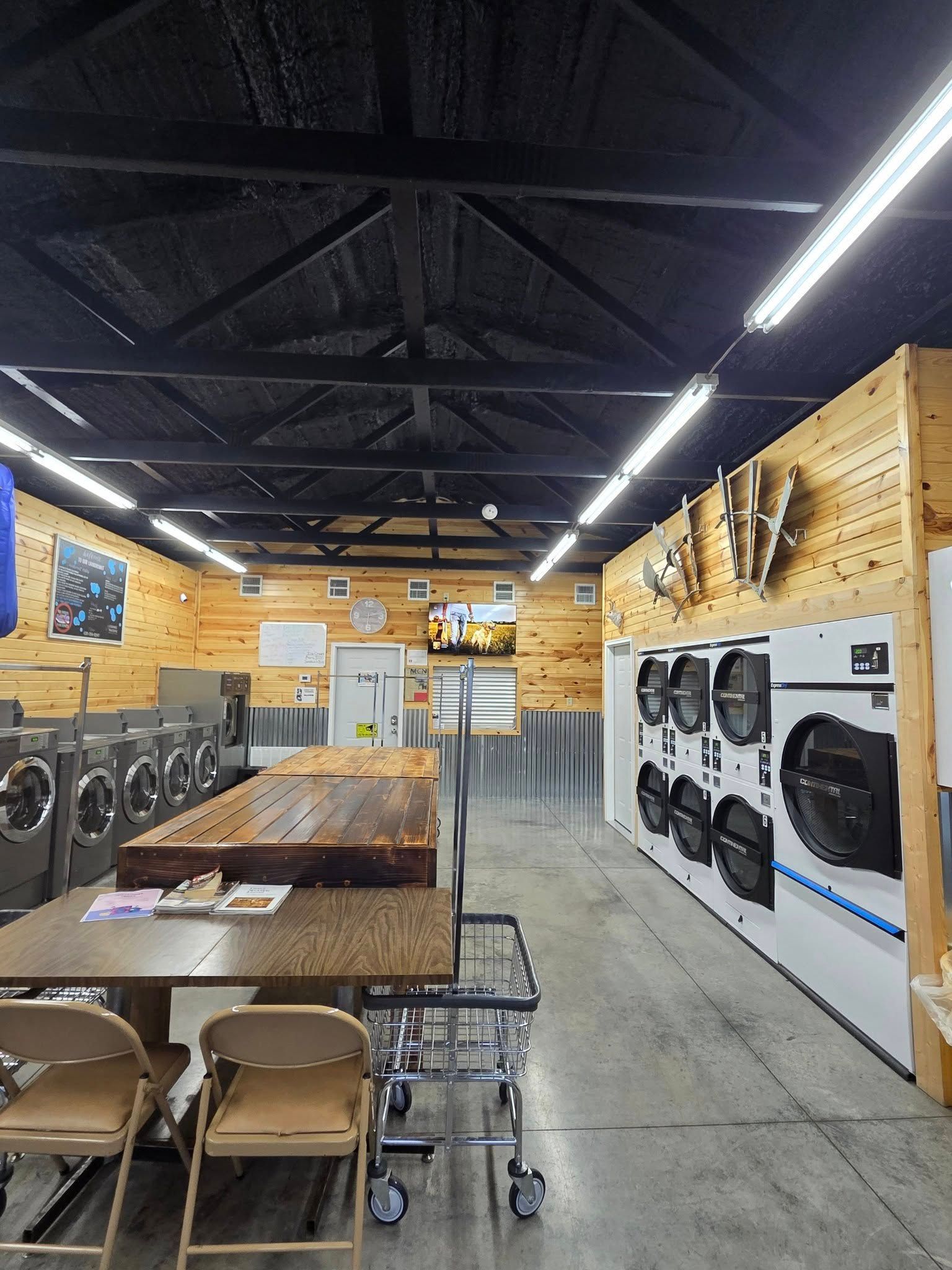Laundry room with rows of washers and dryers, wooden tables, and black ceiling lights under exposed beams.