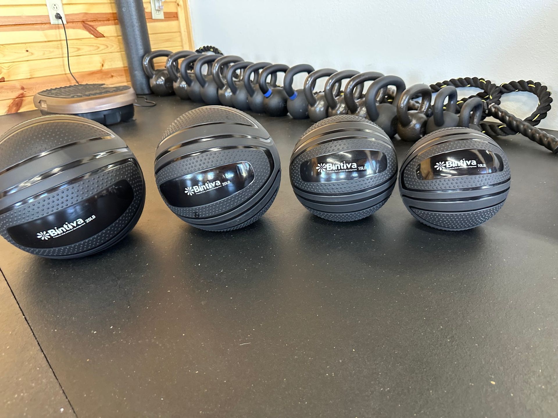 Four black medicine balls on a gym floor with several kettlebells lined up in the background
