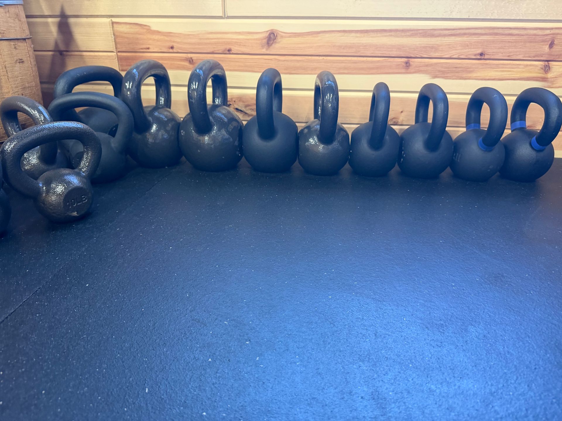 Row of black kettlebells and circular weights on a blue floor against a wooden wall.
