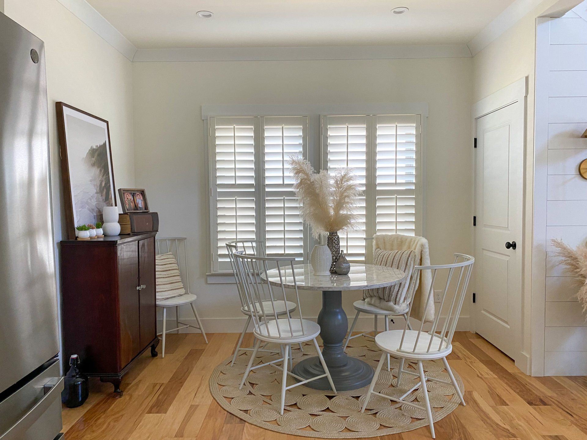 Bright dining room with white chairs, round table, shutters, and a piano along the wall