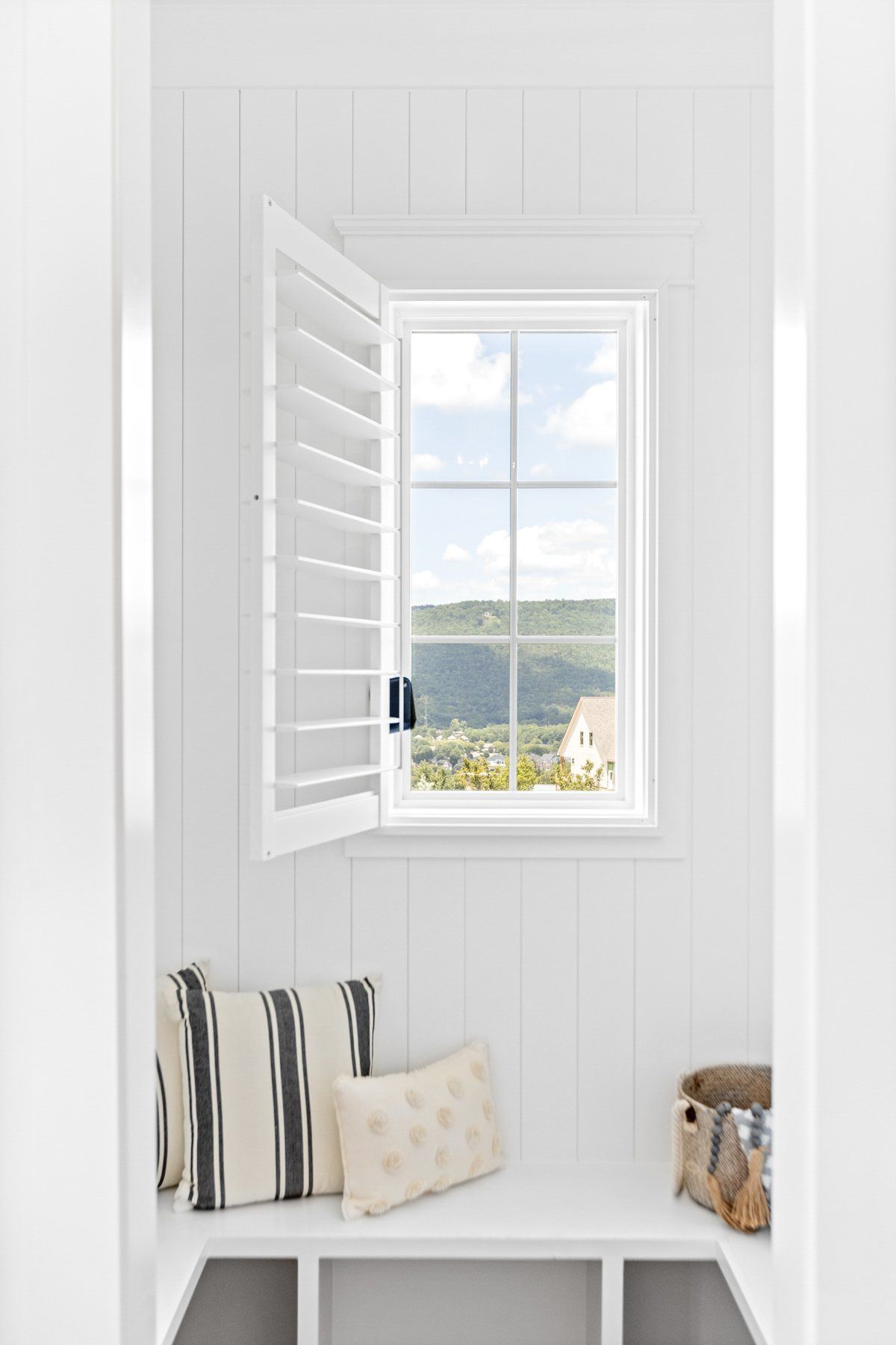 White window nook with shutters, built-in bench, striped pillows, and a lake view outside.