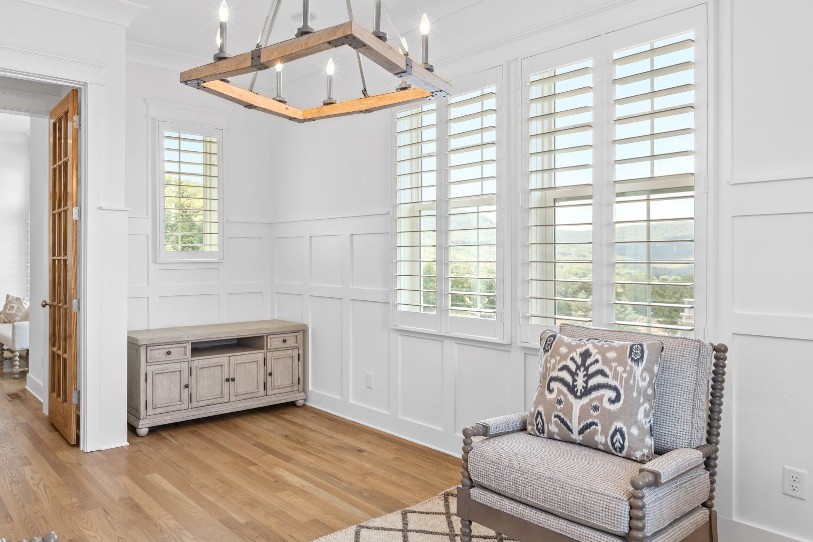 Bright white dining room with wood floors, chandelier, shuttered windows, bench, and patterned armchair