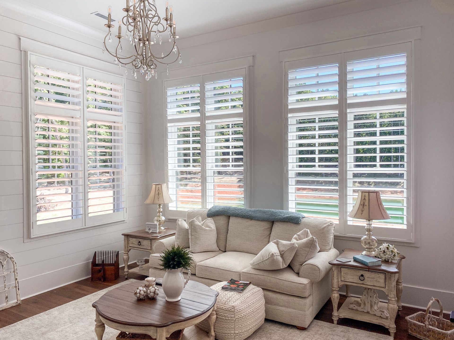 Bright white living room with shuttered windows, cream sofa, round coffee table, and chandelier