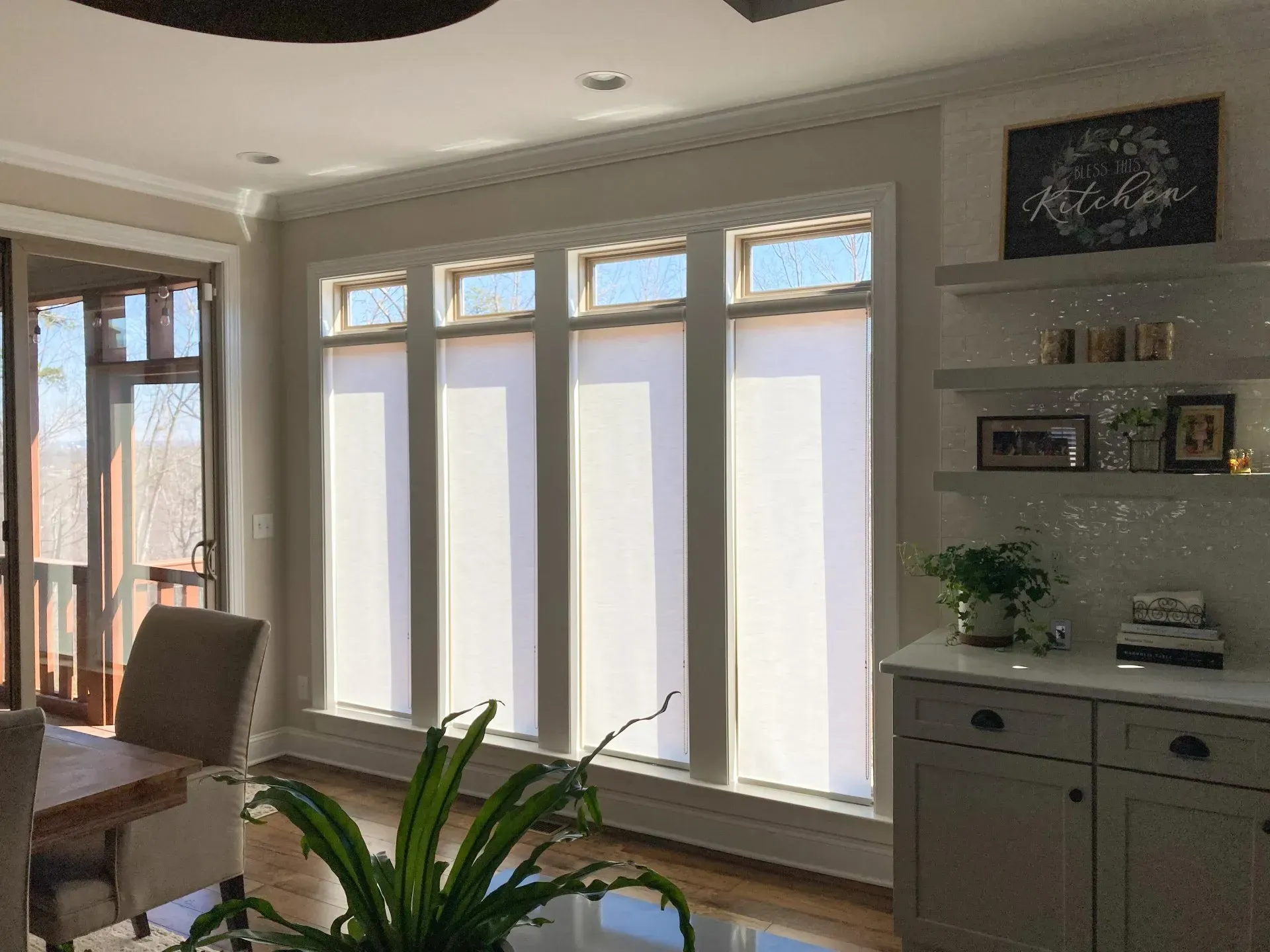 Bright living room with large windows, white blinds, a dining table, cabinet, and a potted plant.