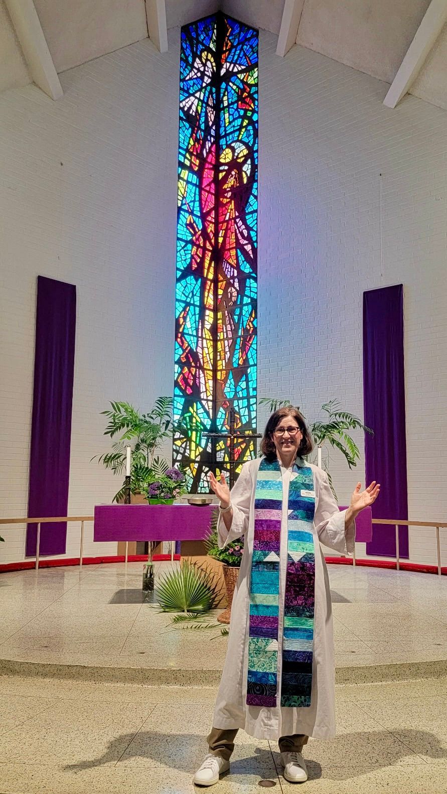 Pastor in a colorful stole stands at a church altar with a tall stained-glass window behind them.