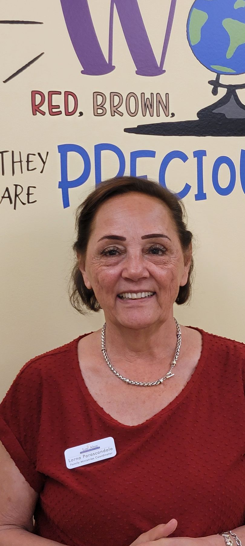 Smiling woman in a red shirt and necklace standing in front of a wall with colorful text.
