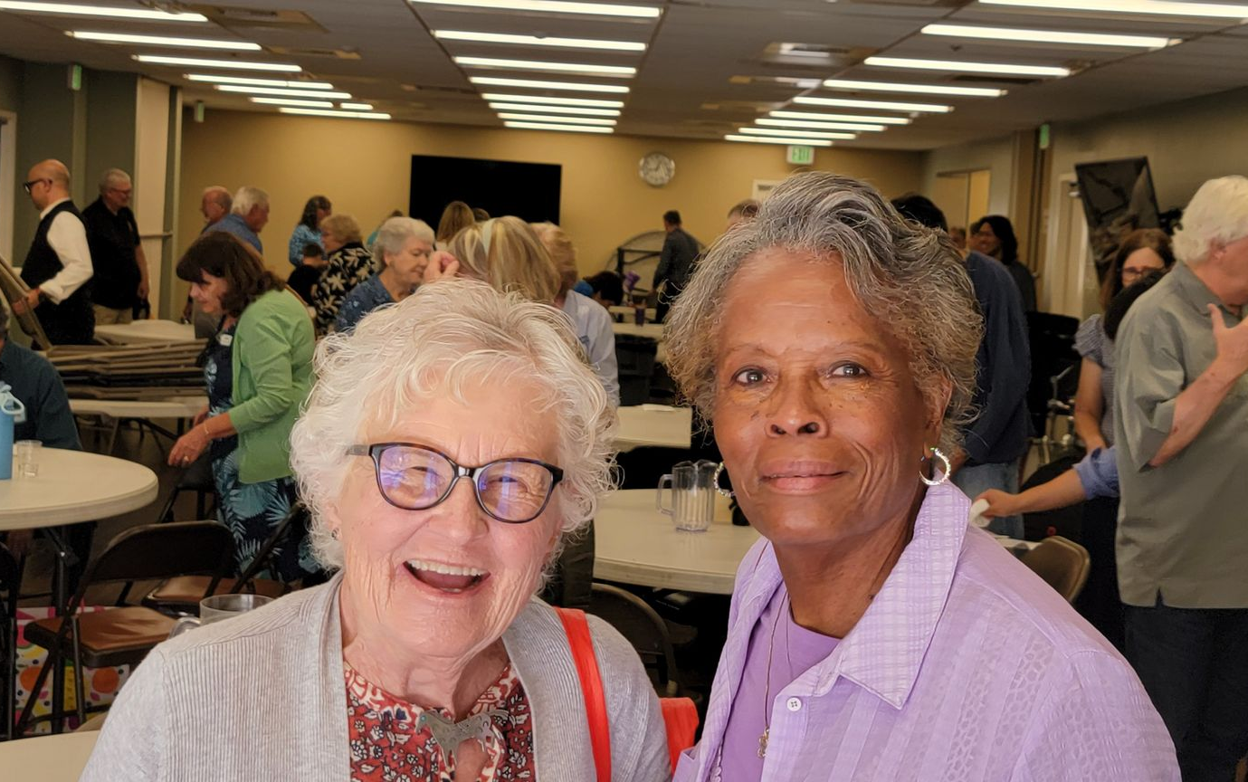 Two women smiling at a crowded indoor gathering with tables and people in the background