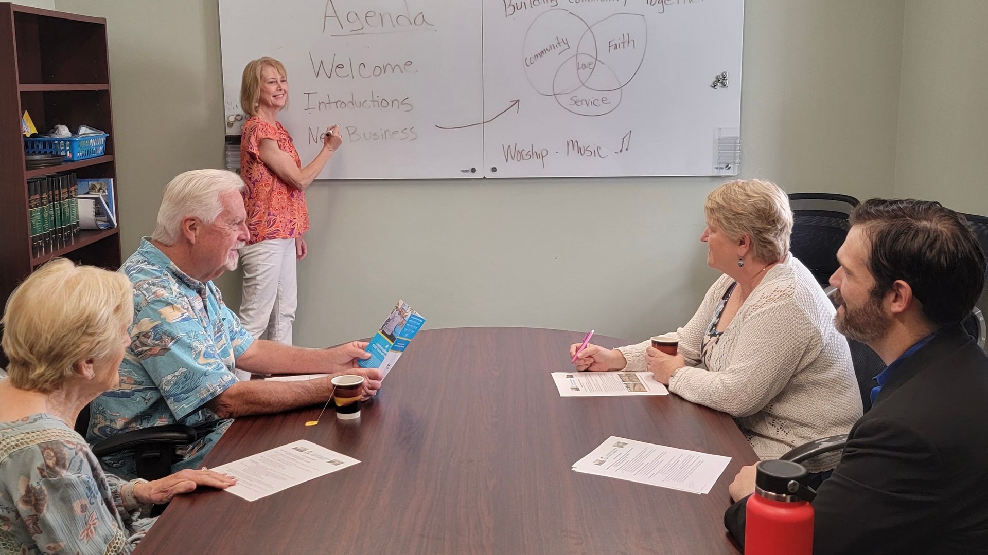 Small meeting room with a woman presenting at a whiteboard while four people sit around a conference table. Small meeting room with a woman presenting at a whiteboard while four people sit around a conference table.