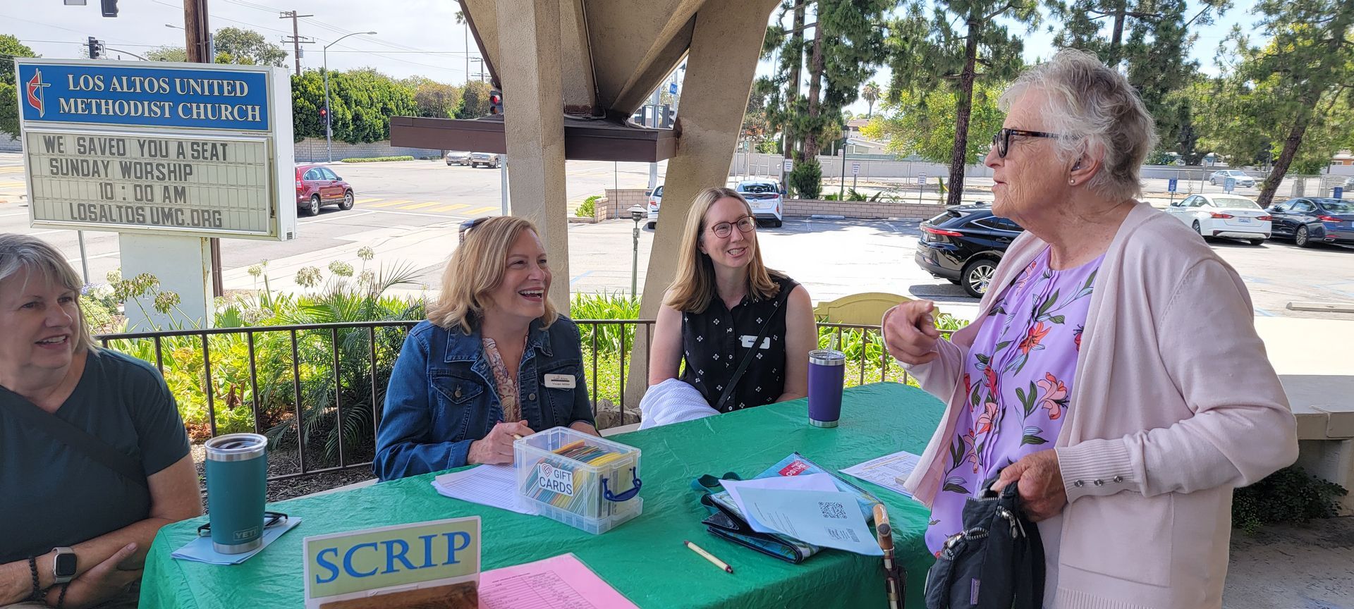 People seated at a green table under an overpass, talking at an outdoor community booth People seated at a green table under an overpass, talking at an outdoor community booth