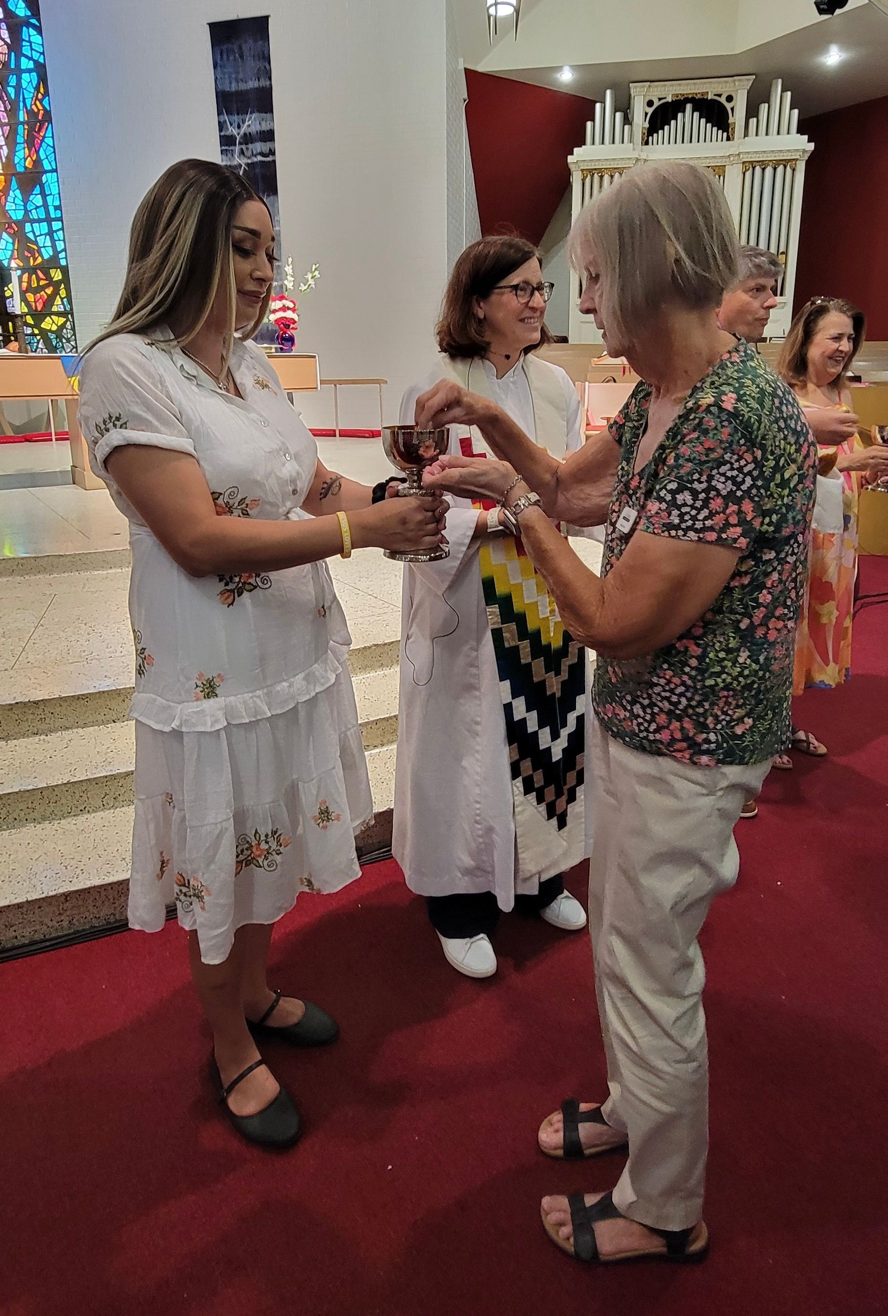 Two women exchanging an item in a church aisle, with others standing nearby and a red carpeted floor.