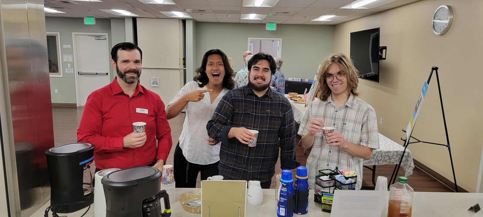 Four people smiling and holding coffee  at a casual indoor gathering with a table of snacks and bottles.