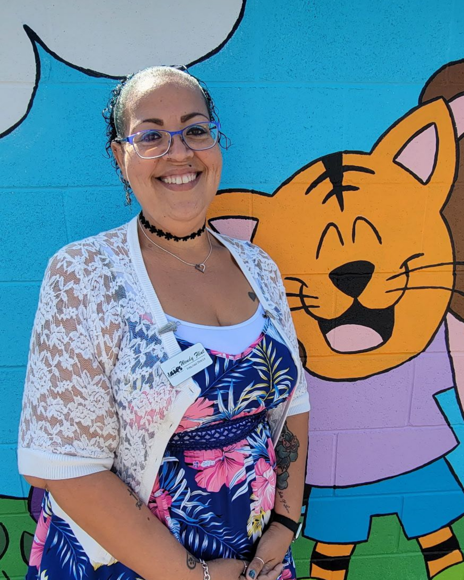 Smiling person in glasses standing before a colorful mural, wearing a white floral cardigan and patterned top