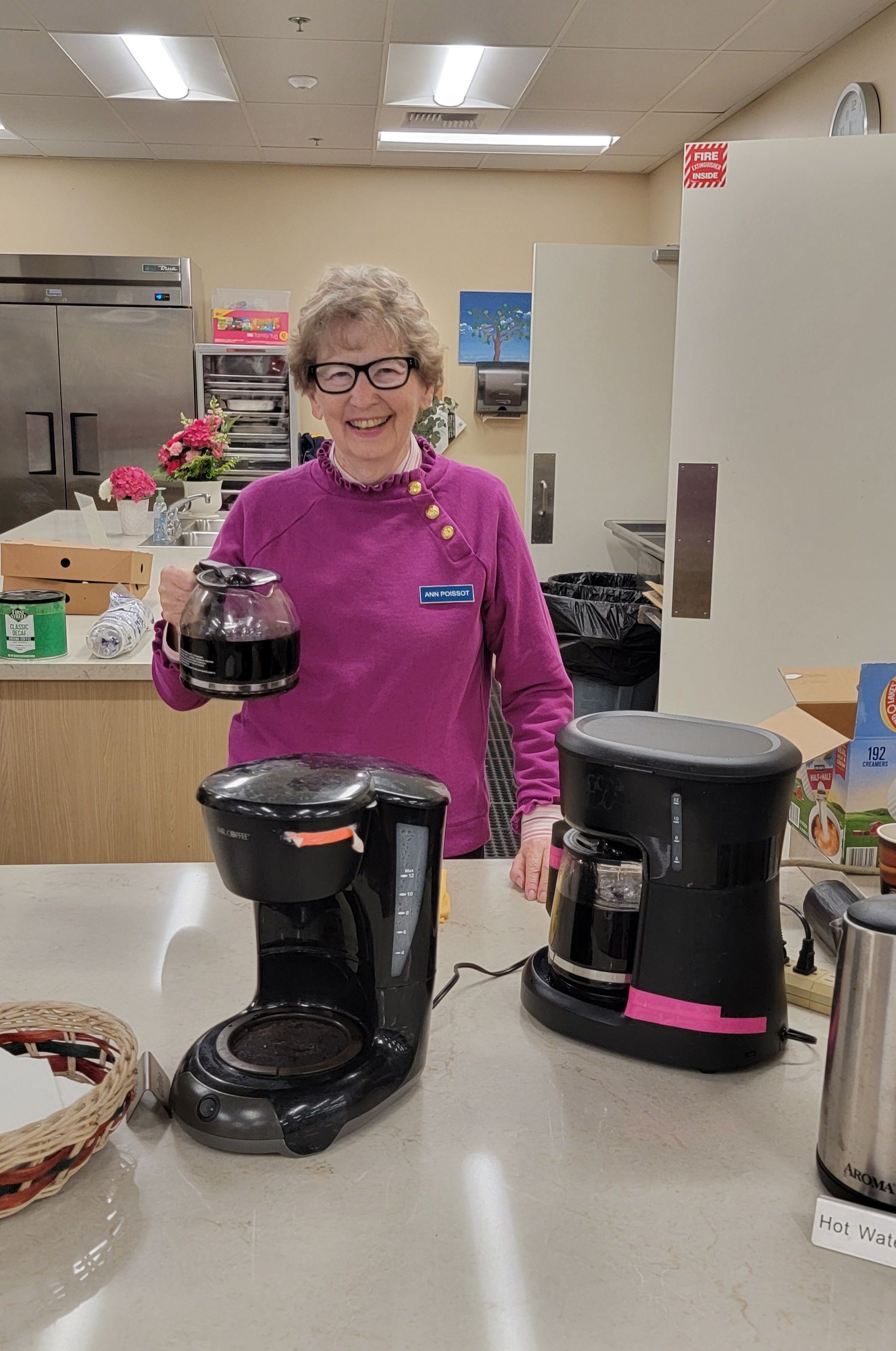 Woman in a kitchen holding a coffee pot beside two black coffee makers on a counter.