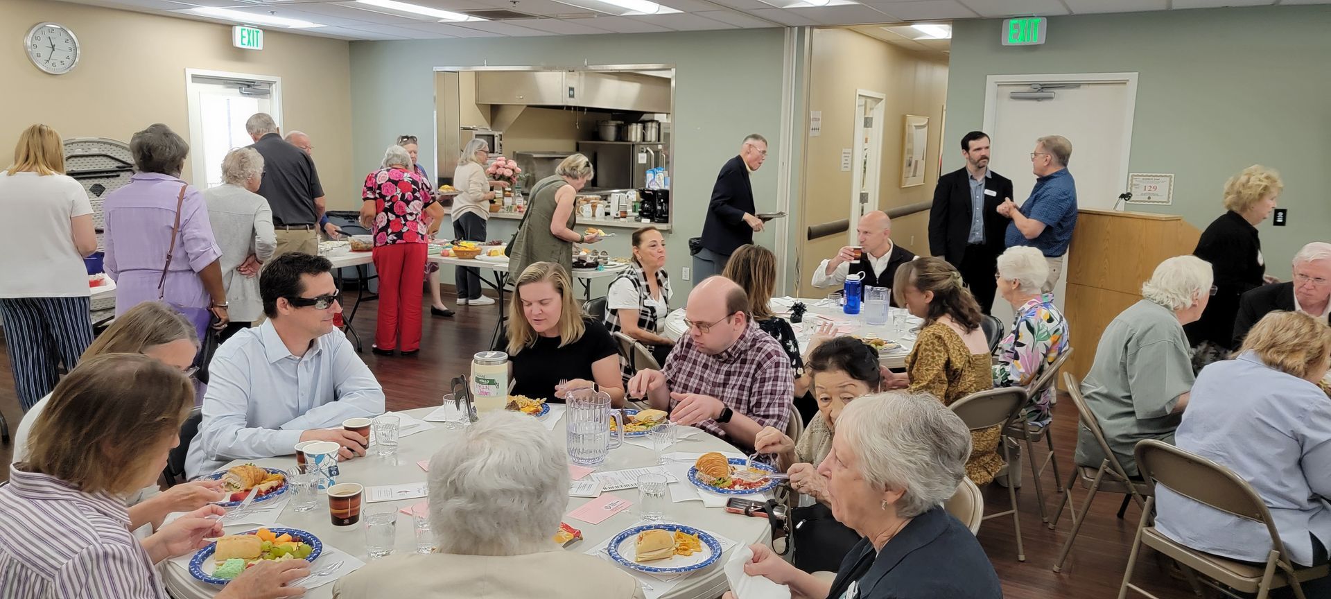 Community room with people seated at tables, eating and talking during a social gathering Community room with people seated at tables, eating and talking during a social gathering