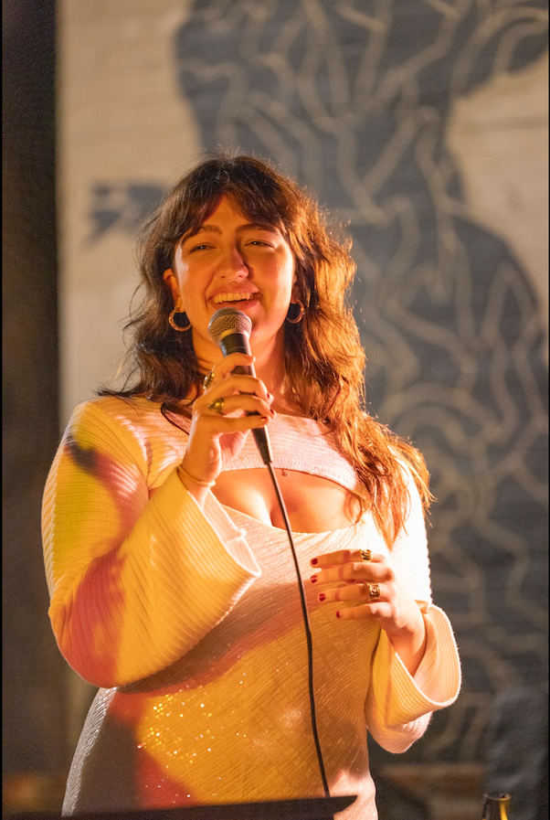 Woman singing into a microphone onstage in a yellow-and-white top under warm lighting