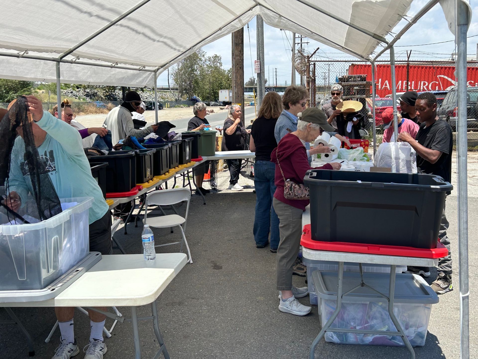 People outdoors under white tents, with tables of food and supplies. People outdoors under white tents, with tables of food and supplies.