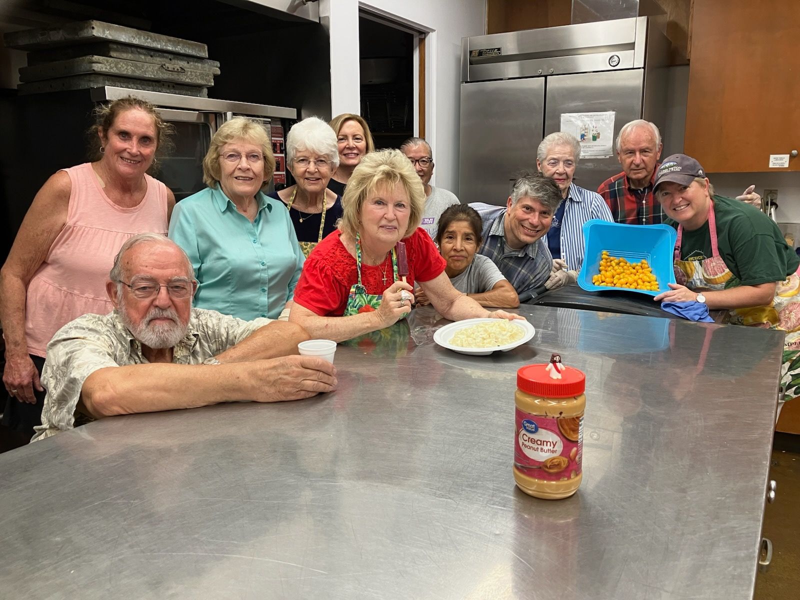 Group gathered around a stainless table in a kitchen, smiling beside a cake and a jar of sauce
