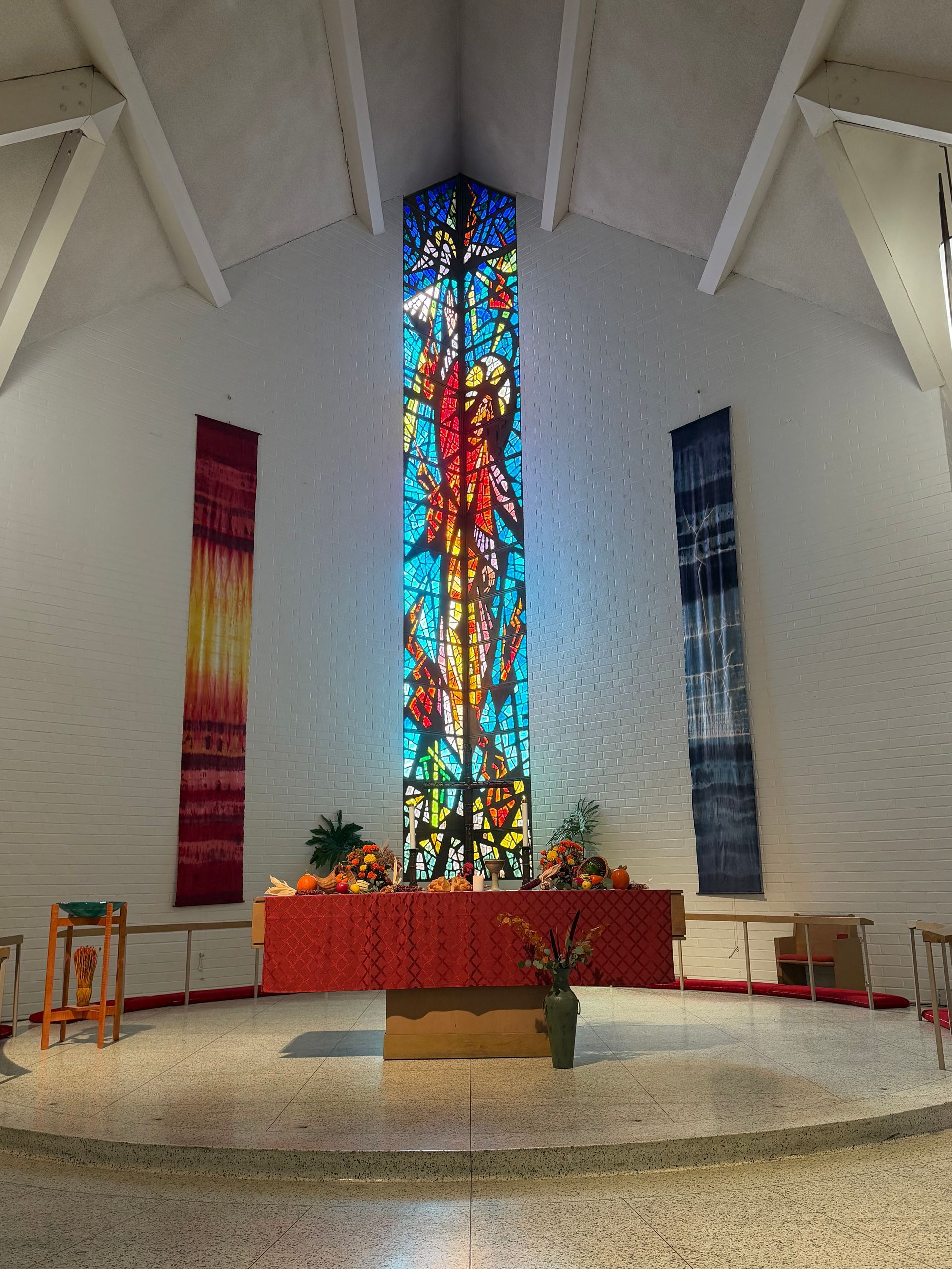 Church altar with tall stained-glass window, red cloth, candles, and flowers in a white sanctuary