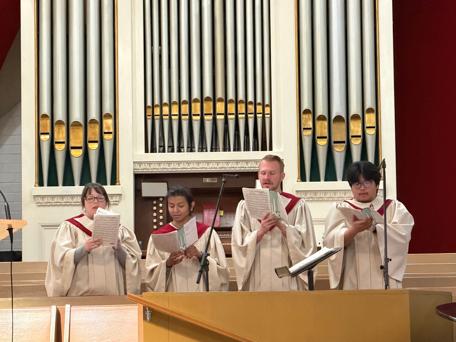 Four people in choir robes holding sheet music and singing in front of organ pipes in a church.