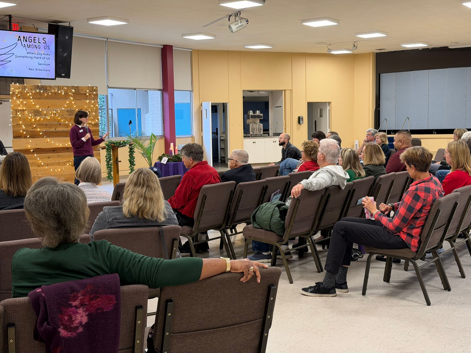 Pastor presenting to seated audience in a community room with projector screens and chairs