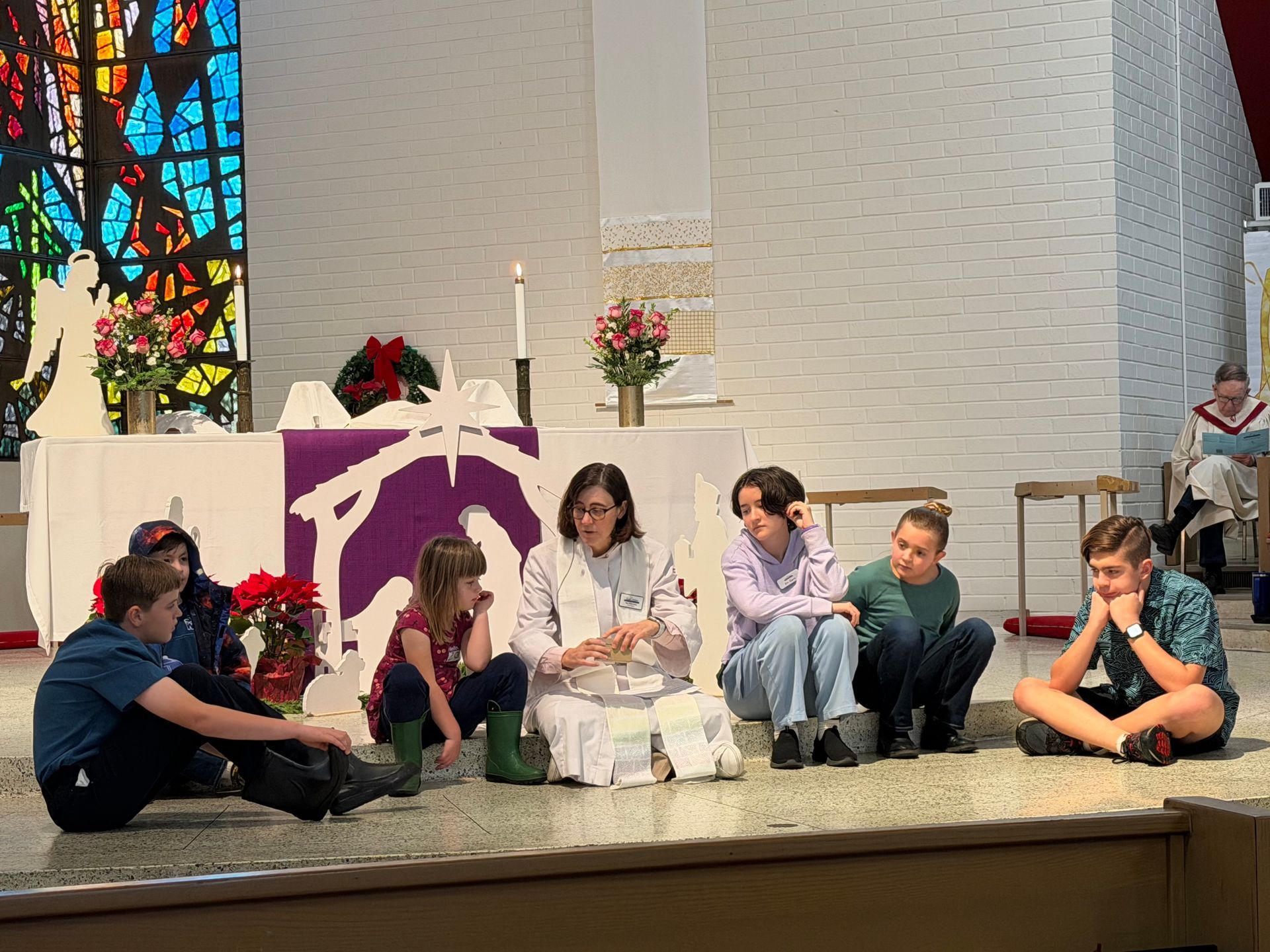 Children seated on a church alter near the pastor, with candles and flowers and a stained glass window in the background