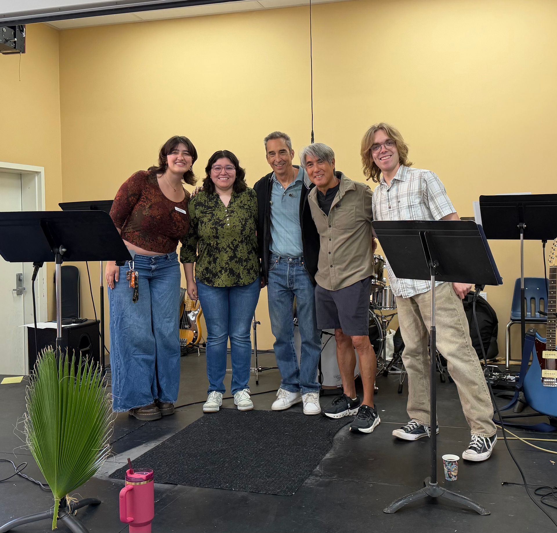 Five people pose smiling on a small stage with music stands and microphone stands. Five people pose smiling on a small stage with music stands and microphone stands.