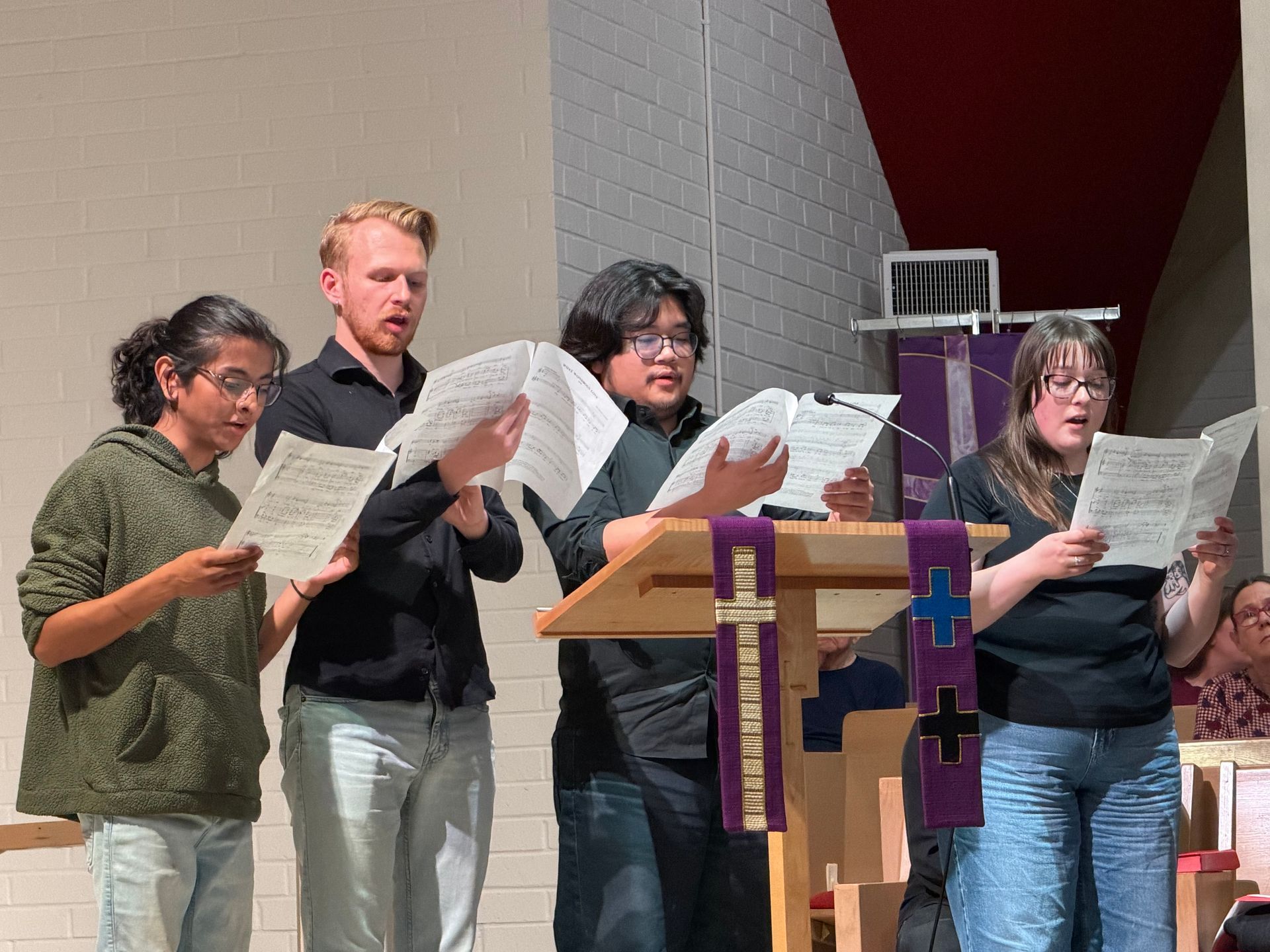 Four people stand behind a podium, holding sheet music and singing in a church. Four people stand behind a podium, holding sheet music and singing in a church.