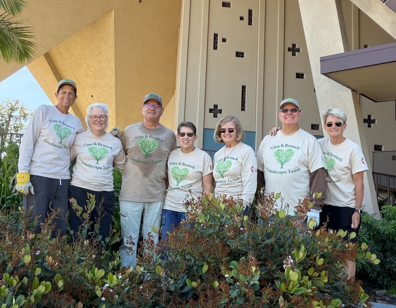 Seven volunteers in matching white T-shirts stand smiling outside a church among shrubs. Seven volunteers in matching white T-shirts stand smiling outside a church among shrubs.