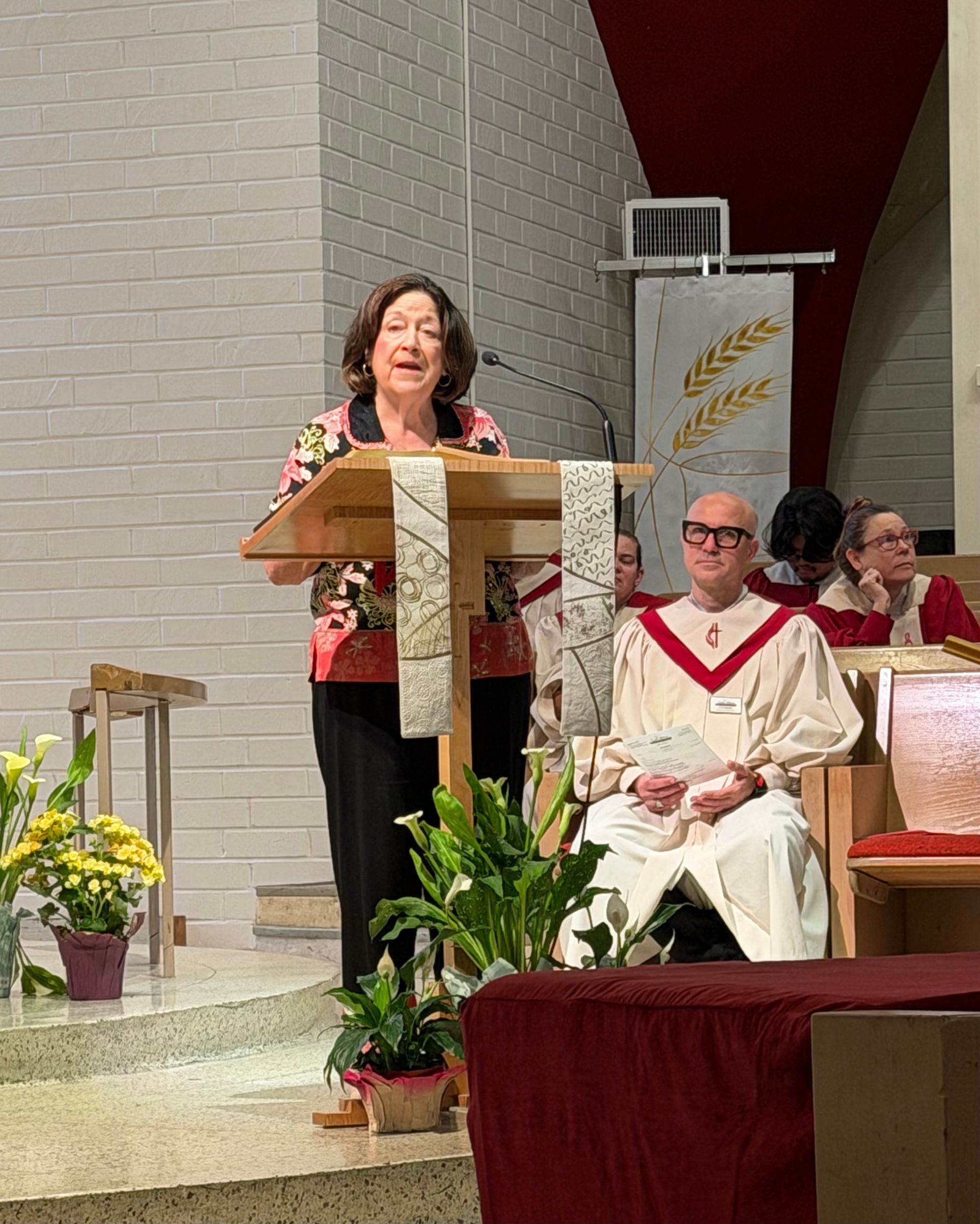 Woman speaking at a church lectern beside a seated choir, with flowers and a red altar cloth in the foreground Woman speaking at a church lectern beside a seated choir, with flowers and a red altar cloth in the foreground