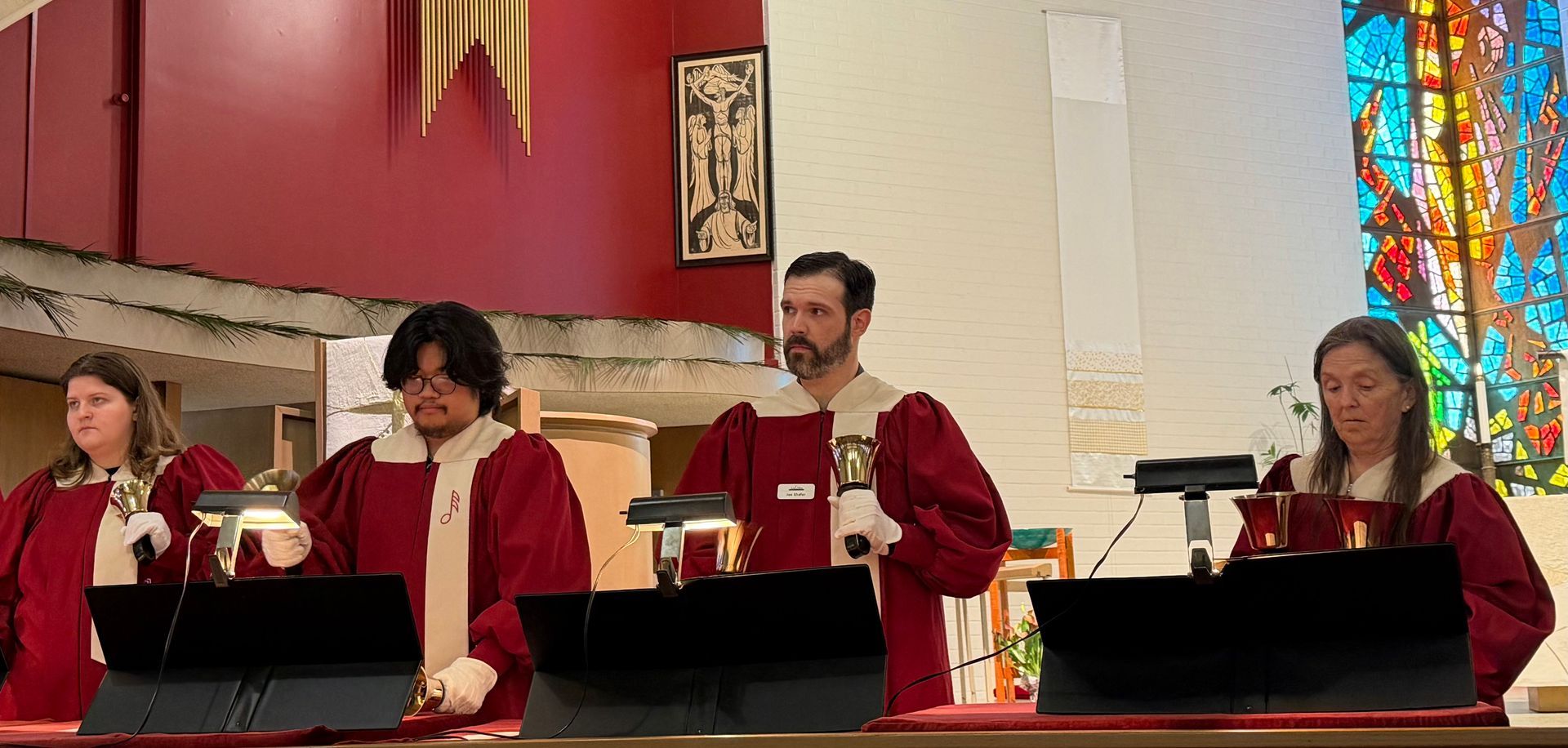 Persons in red robes playing bells at a church altar with stained glass behind them Persons in red robes playing bells at a church altar with stained glass behind them