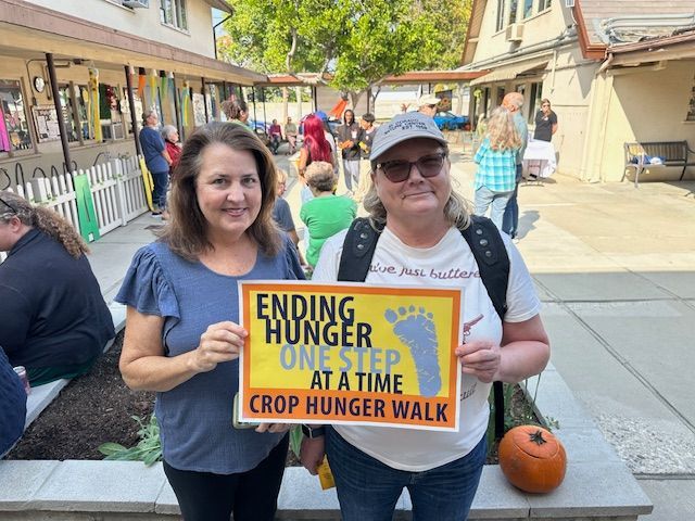 Two women holding an “Ending Hunger One Step at a Time” Crop Hunger Walk sign at an outdoor event Two women holding an “Ending Hunger One Step at a Time” Crop Hunger Walk sign at an outdoor event