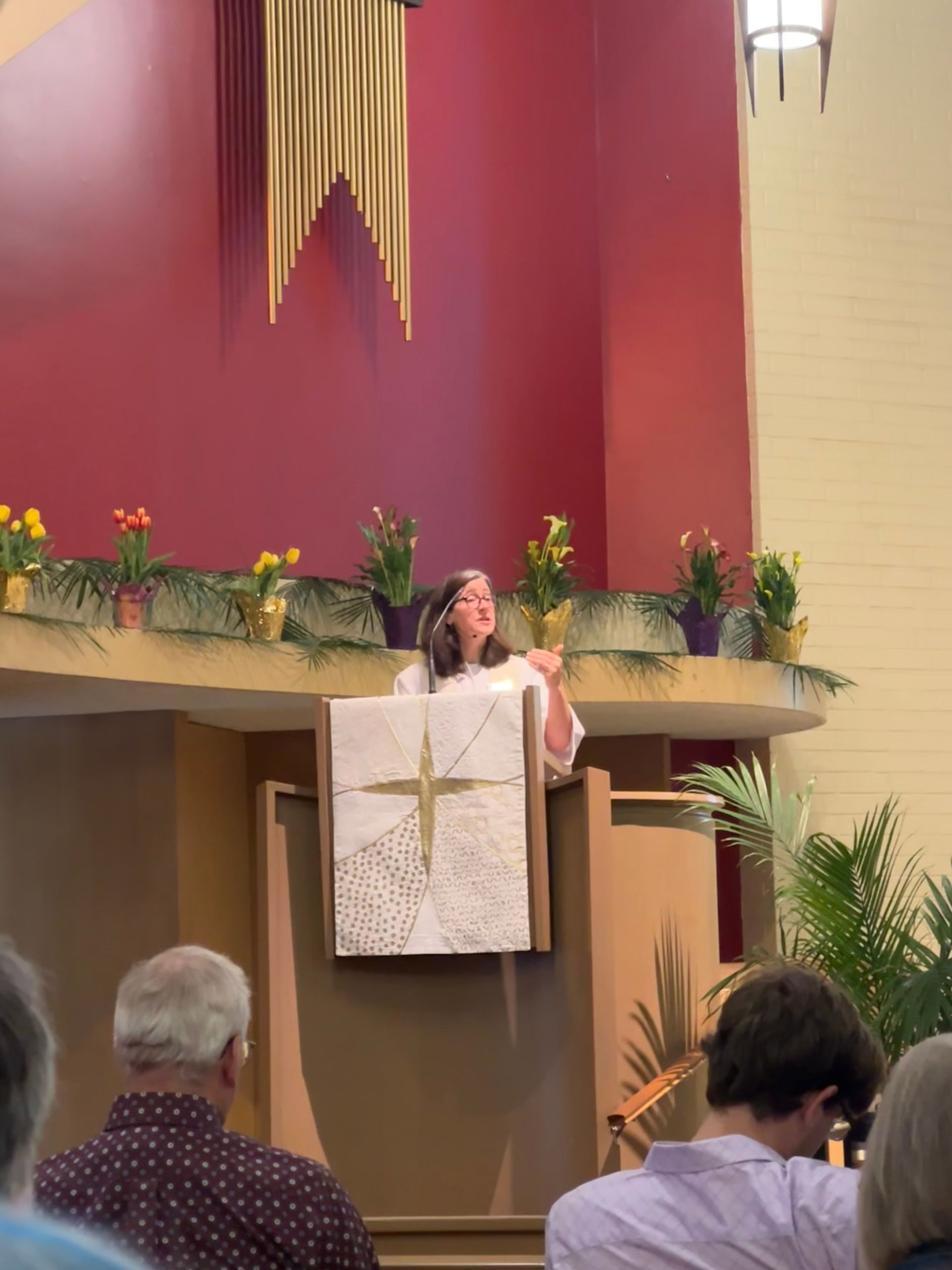 Pastor at church lectern with floral decorations and a cross backdrop, addressing seated attendees