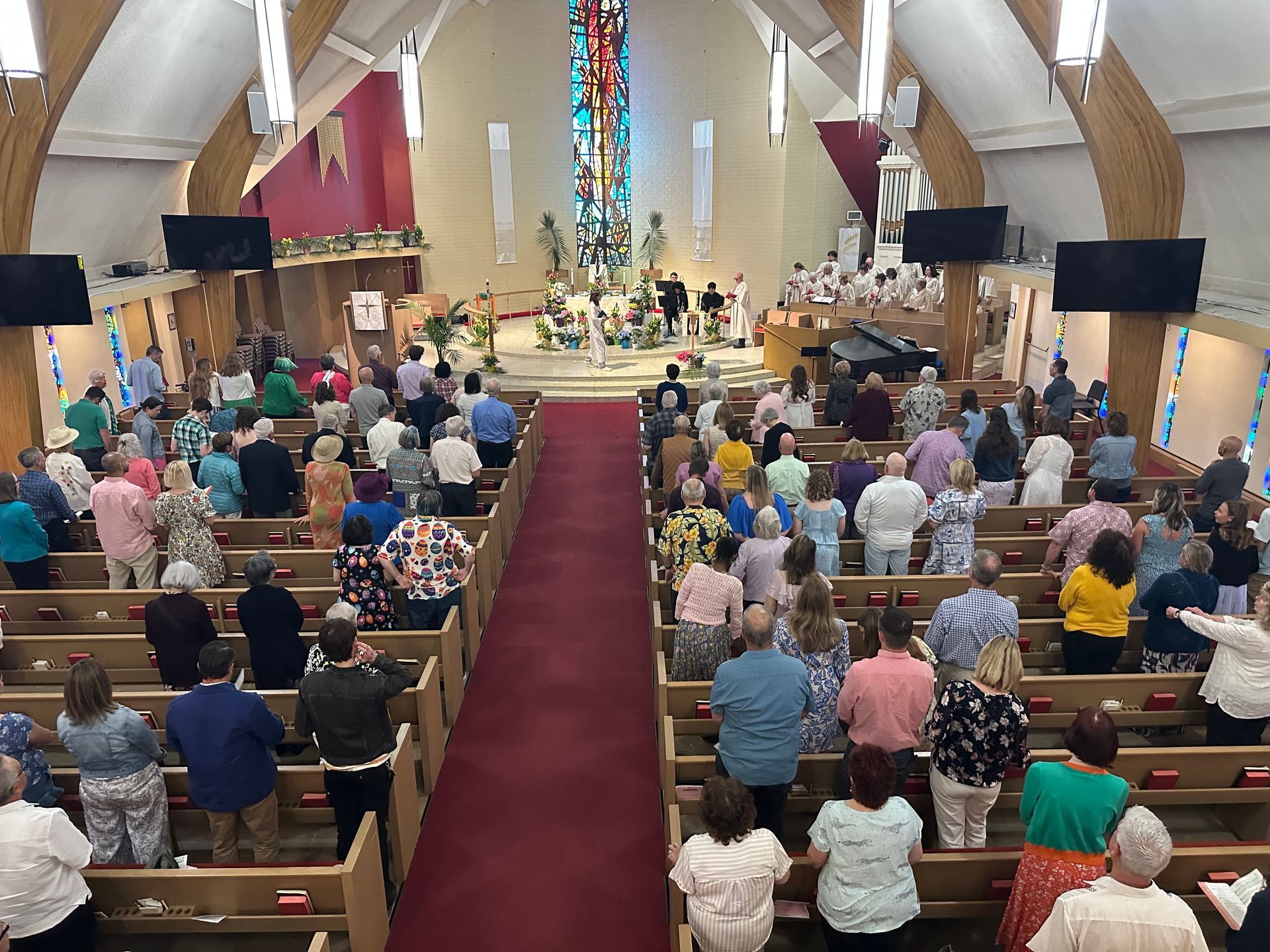 Congregation standing in a church, facing the altar during a service, with stained glass and red carpet aisle