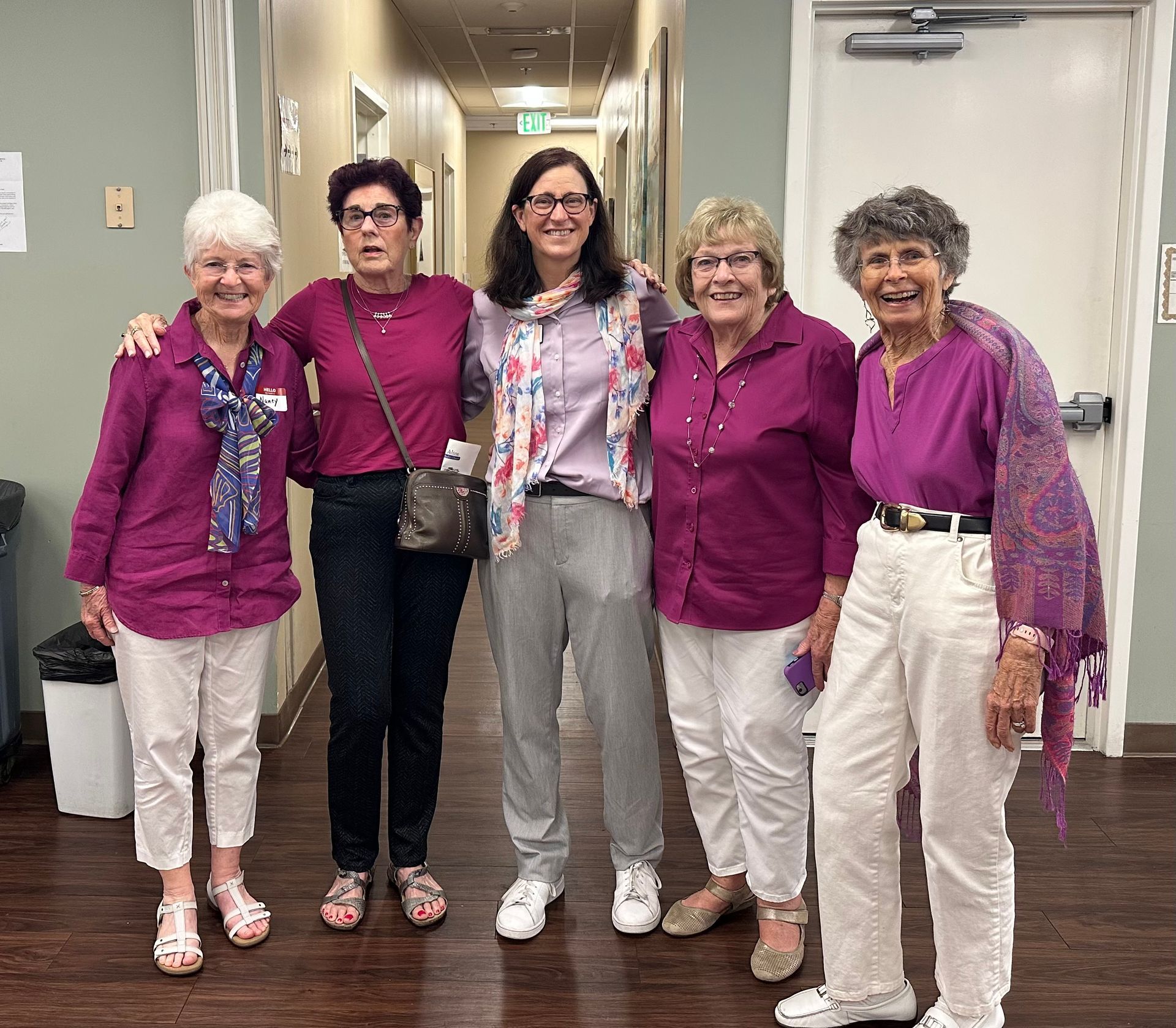 Five women in purple tops standing together in a hallway, smiling at the camera.