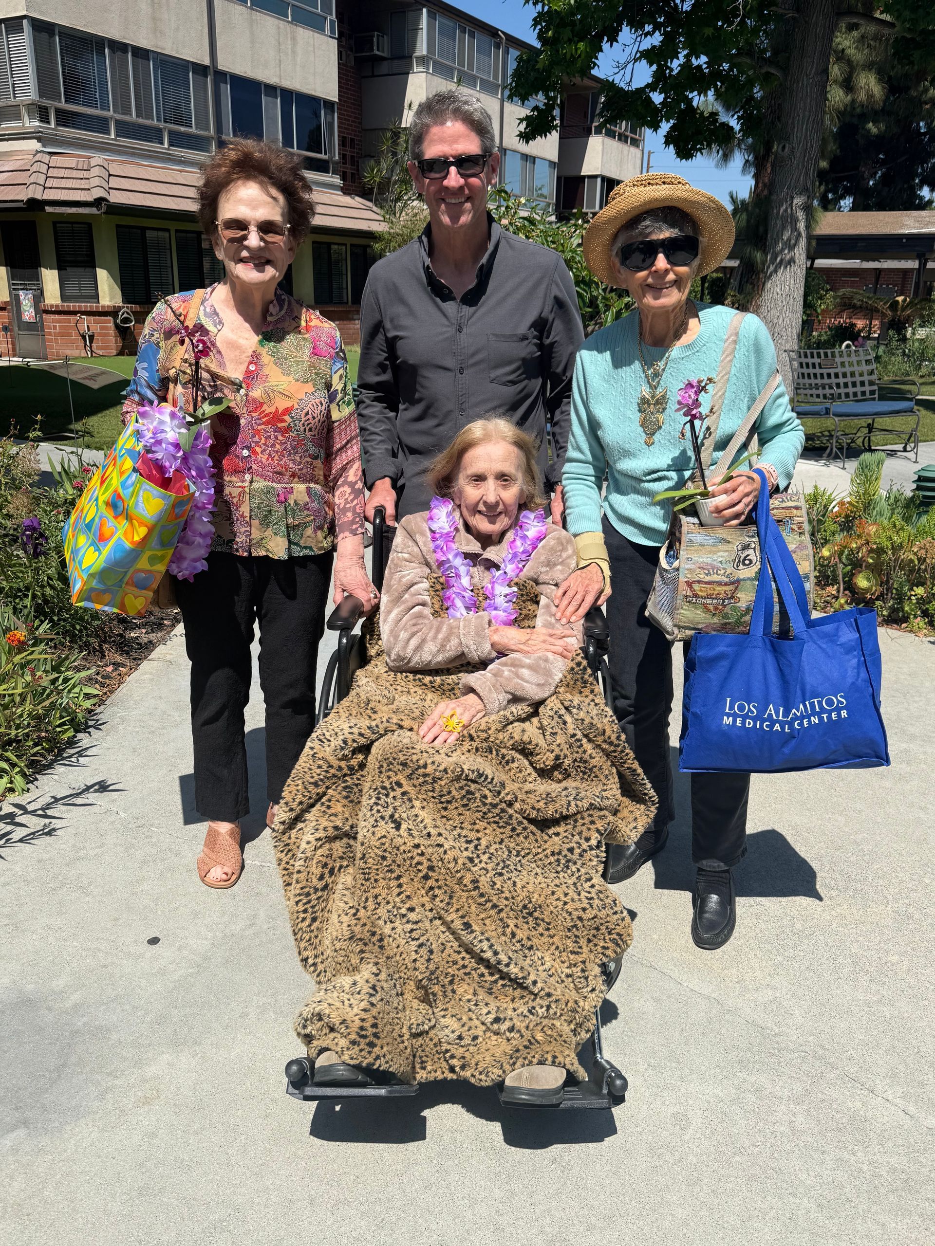 Four people outdoors, one seated, wearing colorful outfits and holding bags, on a sunny walkway. Four people outdoors, one seated, wearing colorful outfits and holding bags, on a sunny walkway.