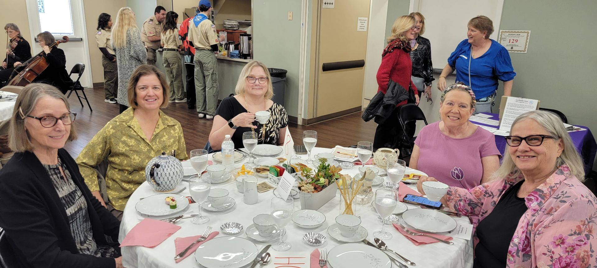 Guests seated around a decorated banquet table in a community hall, smiling and chatting. Guests seated around a decorated banquet table in a community hall, smiling and chatting.