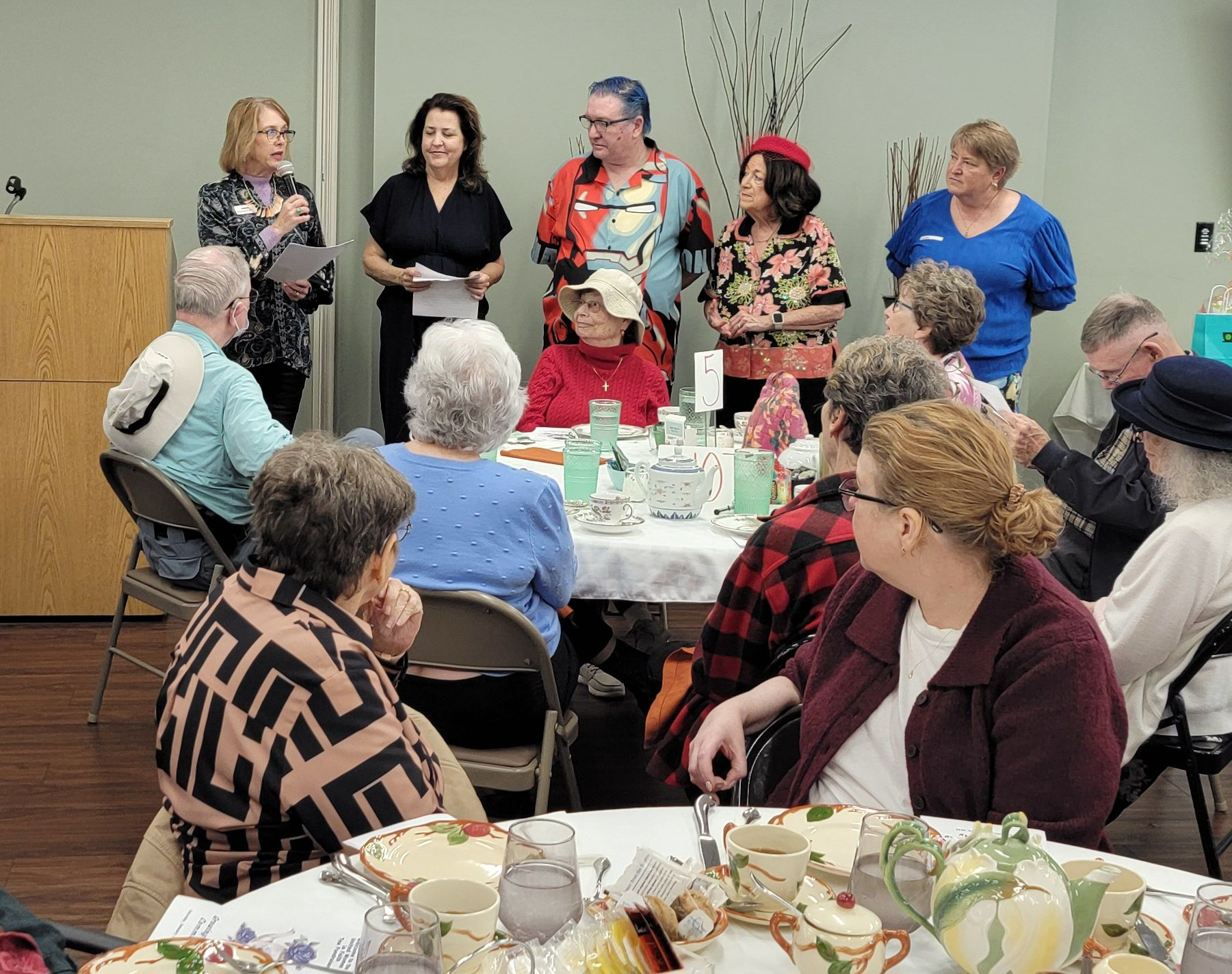 People gathered around tables in a bright room, listening to a speaker at the front. People gathered around tables in a bright room, listening to a speaker at the front.