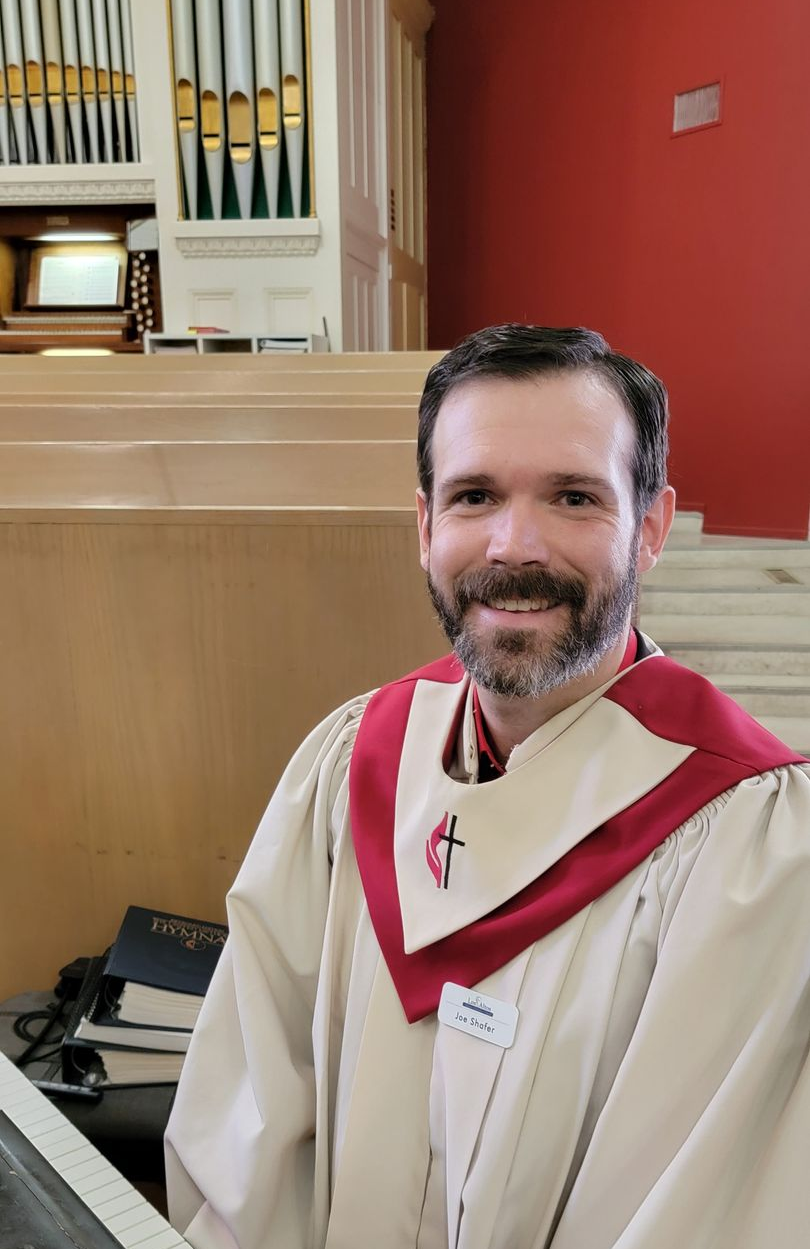 Smiling man in white and red choir robe standing in a church near an organ and wooden pews