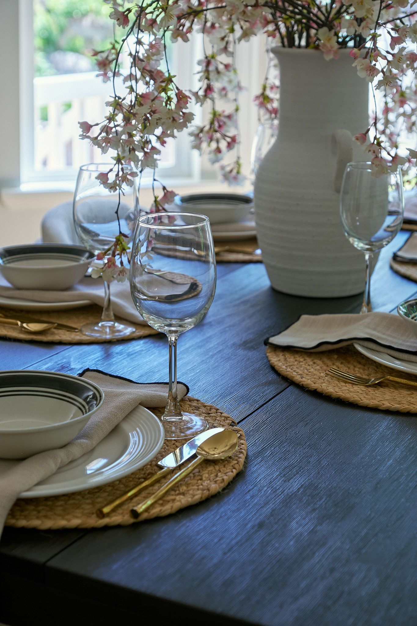 A rustic dining table set with woven placemats, white bowls, gold cutlery, and wine glasses, with a floral centerpiece.