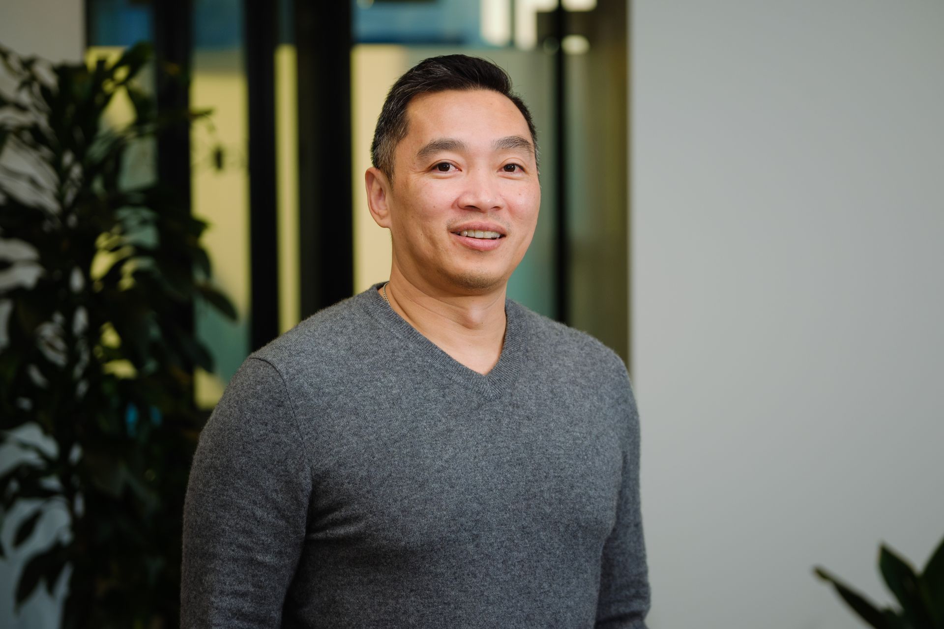 A person wearing a grey v-neck sweater smiles while standing in an office setting with a plant in the background.