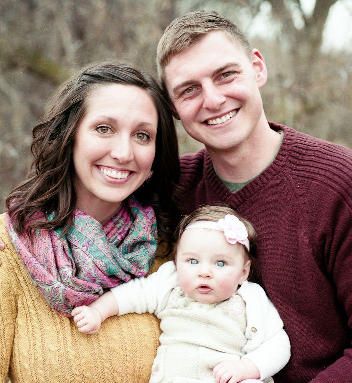 A smiling couple holds a baby with a pink headband, posing outdoors in a yellow sweater and maroon sweater.