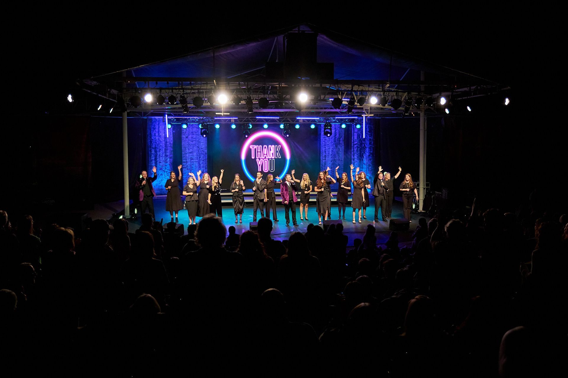 A group of performers standing on a stage under bright lights, facing an audience, with a large