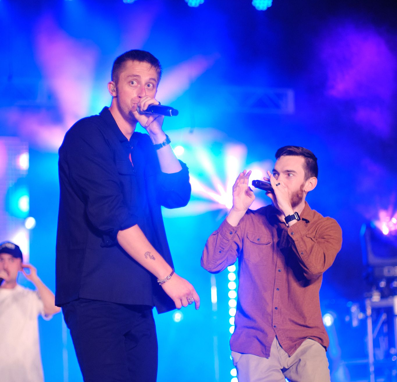 Two performers on a stage with bright blue lighting, singing into microphones in front of a hazy background.