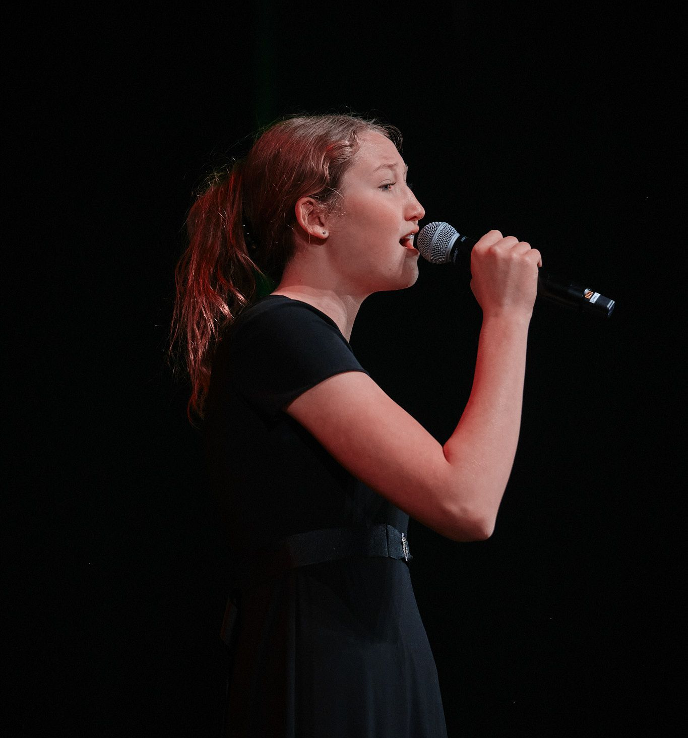A person in a black outfit singing into a microphone on a dark stage.