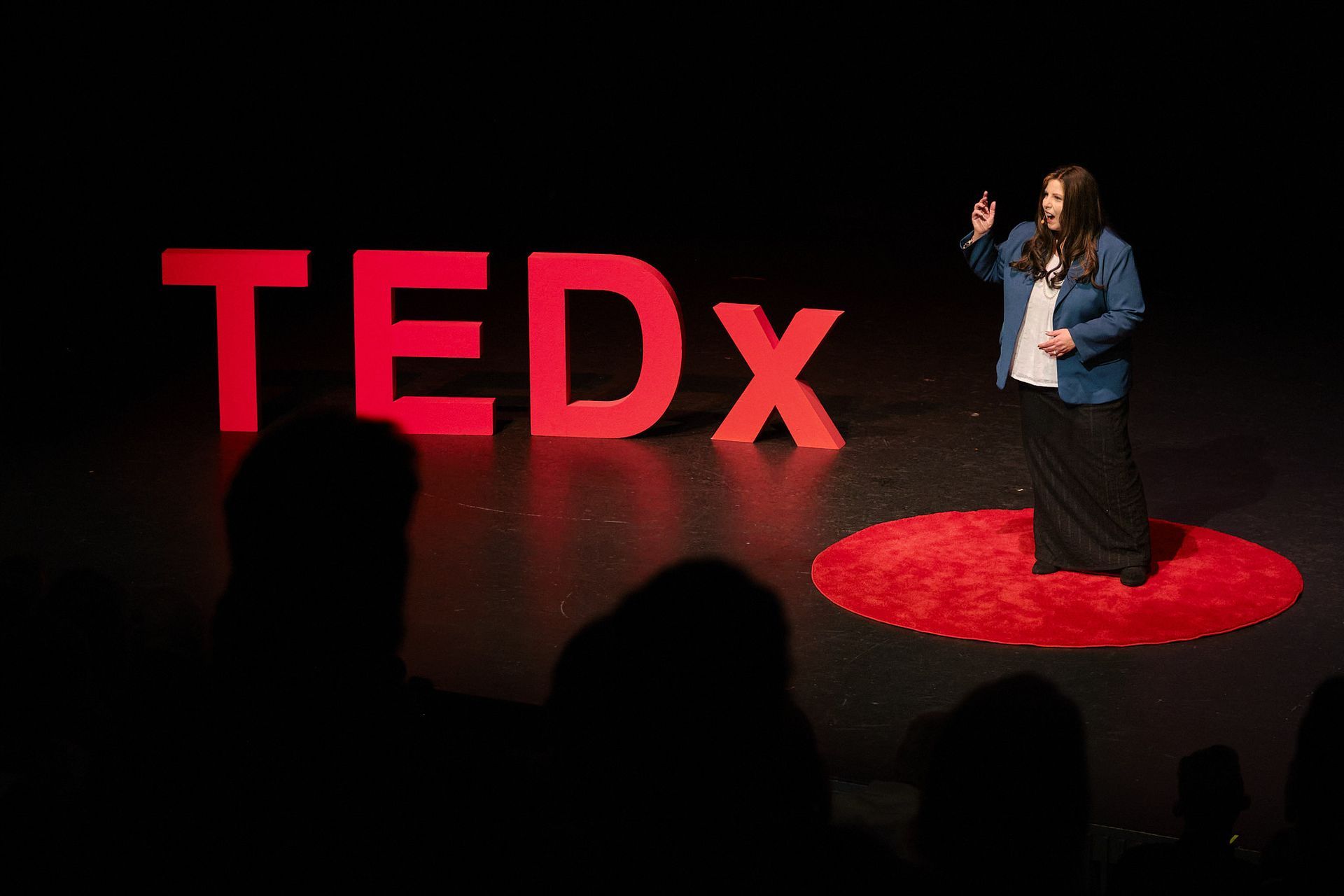 A person gestures while speaking on a stage next to a large red