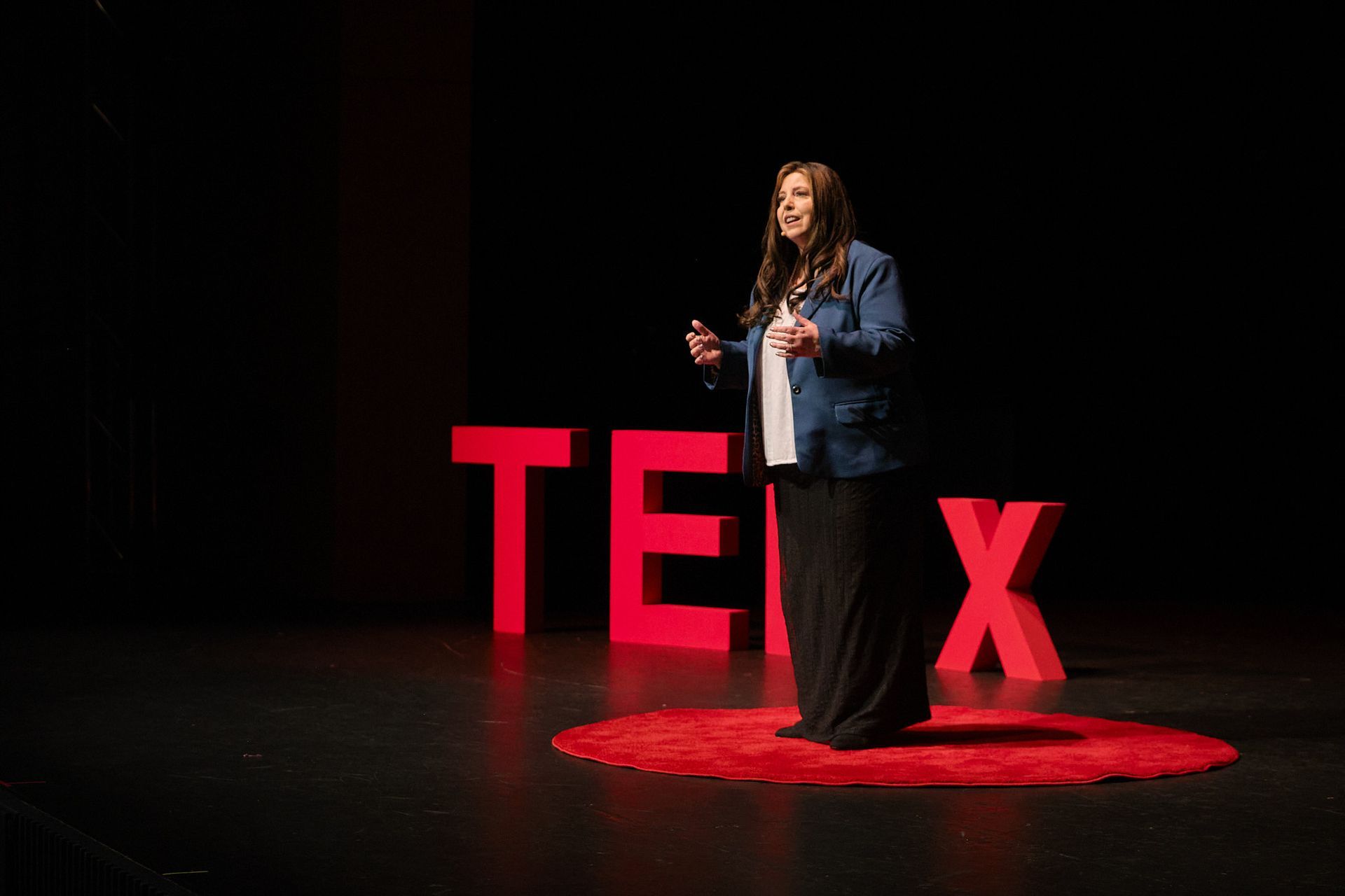 A speaker stands on a circular red rug on a stage in front of large, red