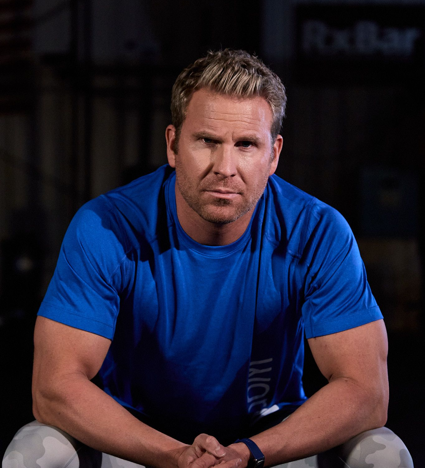 A person with blonde hair in a blue athletic shirt sits with hands clasped, looking forward in a dark gym setting.