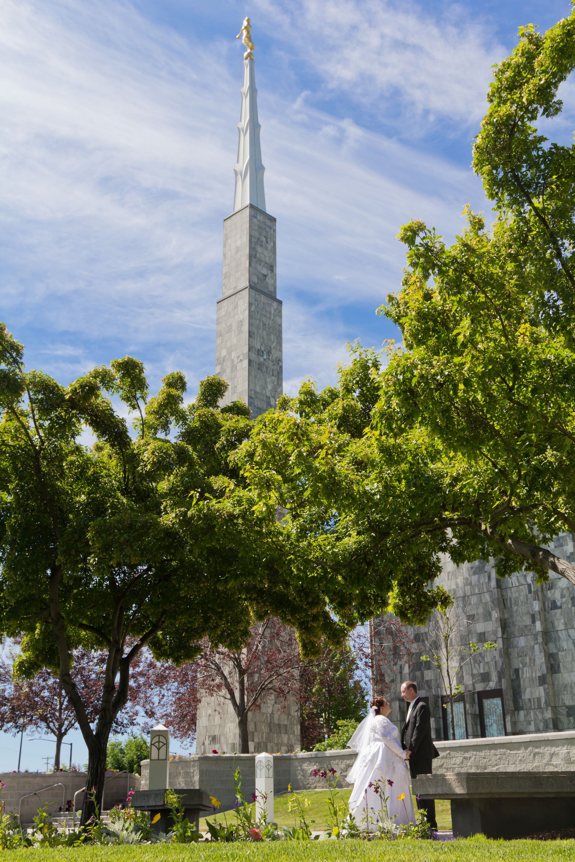 A bride and groom stand outdoors near a tall, stone church spire surrounded by lush green trees under a blue sky.